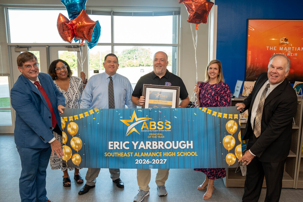 Principal Eric Yarbrough holds his framed award while standing with district leaders behind a blue banner that reads "ABSS Principal of the Year Eric Yarbrough Southeast Alamance High School 2026-2027."