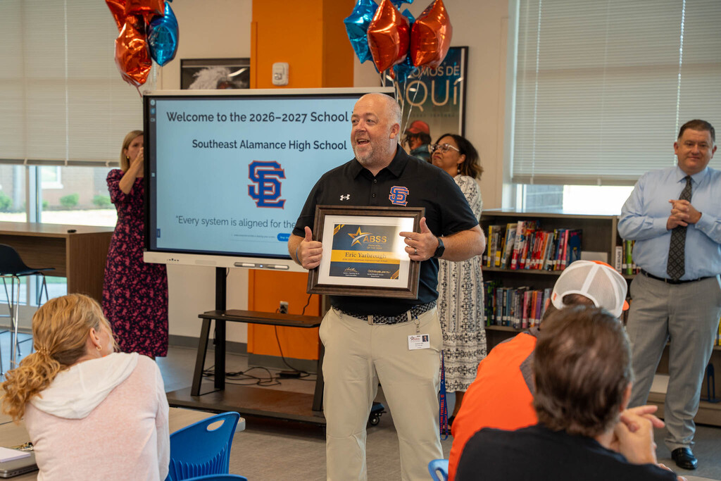 Principal Eric Yarbrough speaks to staff while gesturing toward his framed award certificate in front of a digital display board.