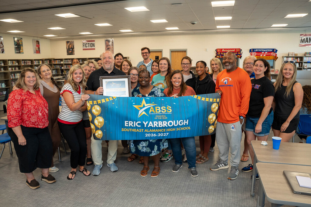 Principal Eric Yarbrough and high school staff gather for a group photo in the media center behind a blue "ABSS Principal of the Year" banner.