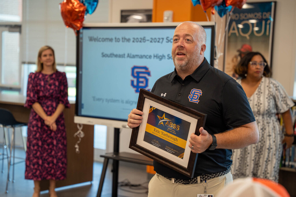Principal Eric Yarbrough holds his framed "ABSS Principal of the Year" certificate in front of a monitor showing the Southeast Alamance High School logo.