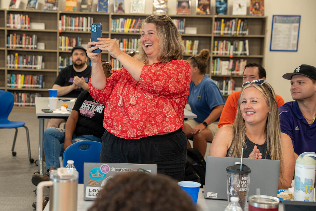A smiling woman in a red patterned top stands up in a school media center to take a smartphone photo during a staff event.