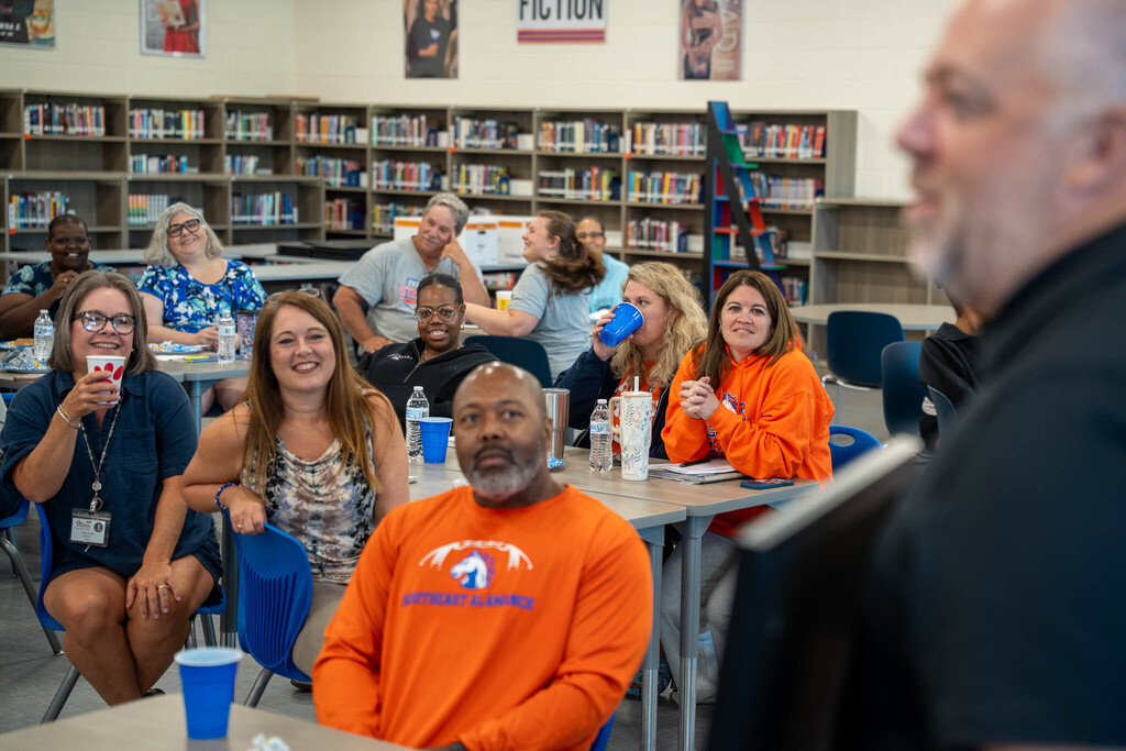 High school staff members sit at library tables, listening attentively and smiling during a presentation.