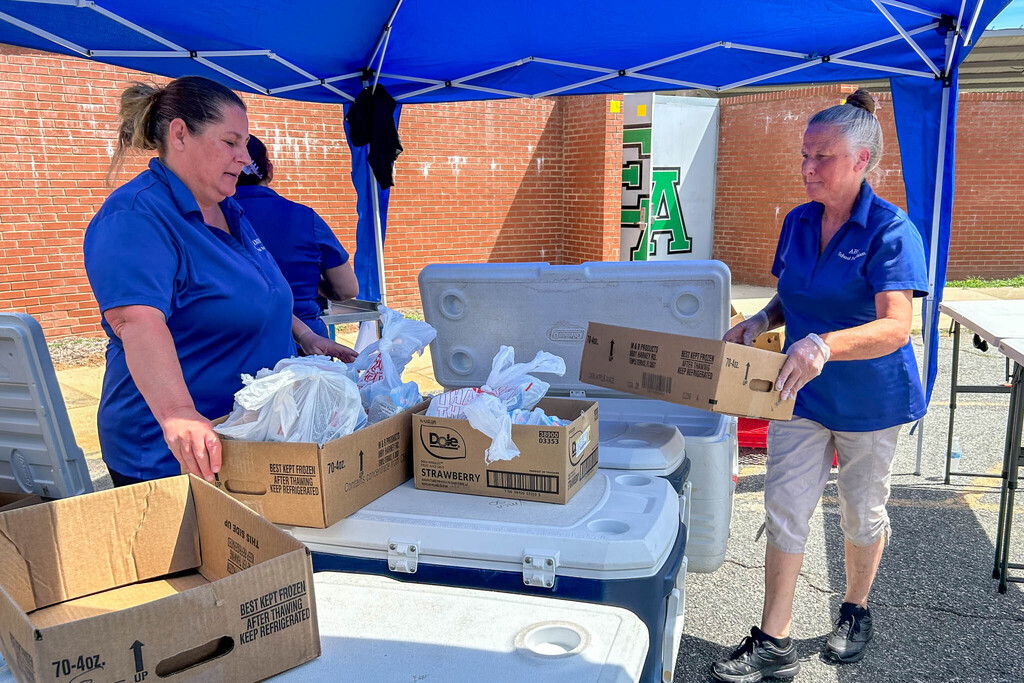 School Nutrition staff wearing blue polo shirts work diligently to distribute bags of meals to families lined up in vehicles in a school parking lot