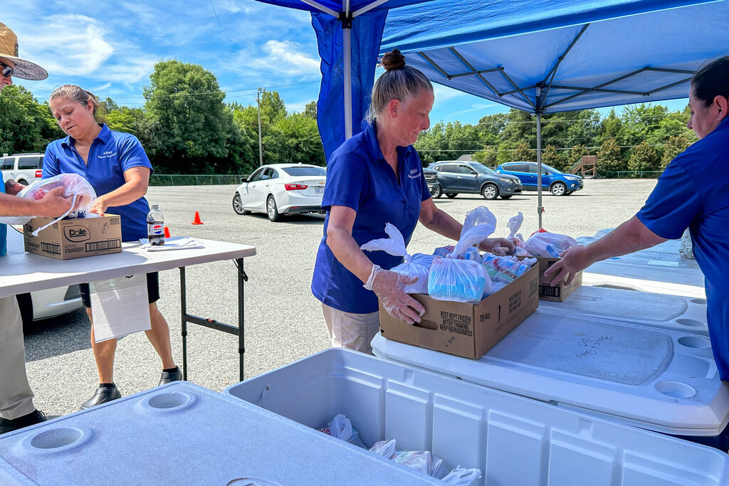 School Nutrition staff wearing blue polo shirts work diligently to distribute bags of meals to families lined up in vehicles in a school parking lot