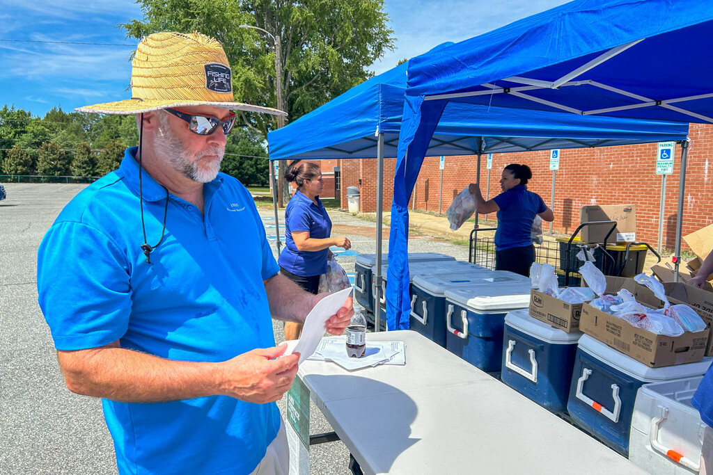 School Nutrition staff wearing blue polo shirts work diligently to distribute bags of meals to families lined up in vehicles in a school parking lot. A man with a brimmed hat looks on a paper list.