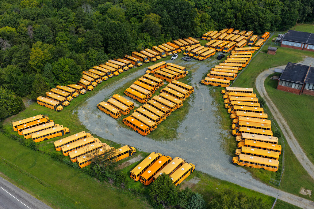 A aerial drone shot of around 50 school buses parked in a grassy area with a gravel road going in the middle in a circle