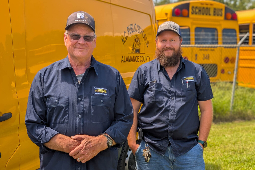 Two bus mechanics stand in front of a yellow NC Public Schools Alamance County van with yellow school buses further in the background