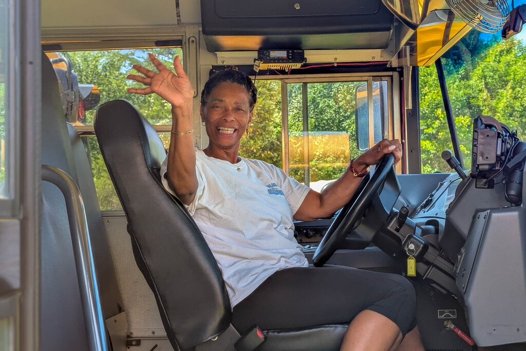A school bus driver wearing a white shirt waves enthusiastically while seated behind the wheel.