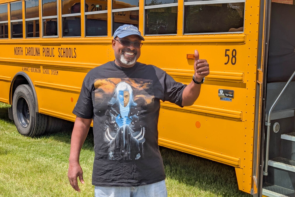 A bus driver wearing a black Prince t-shirt and a blue hat gives an enthusiastic thumbs up beside a yellow school bus