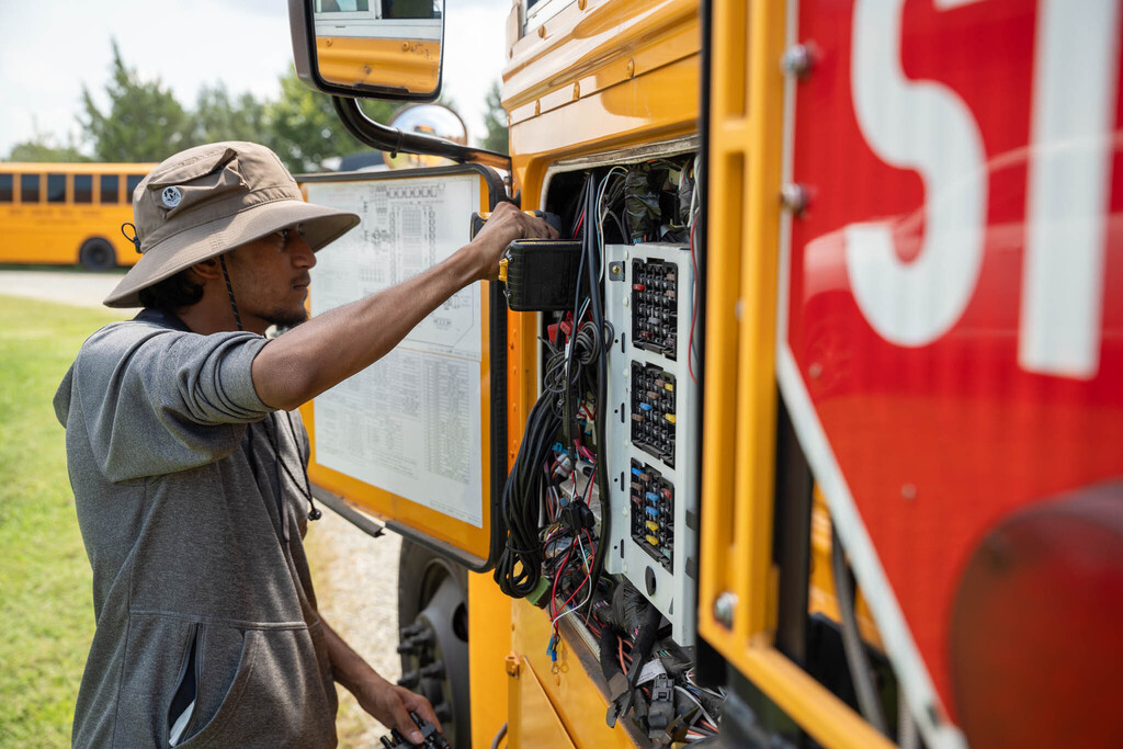 A bus mechanic wearing a sun hat uses a drill on a component inside the fusebox area on the side of a school bus