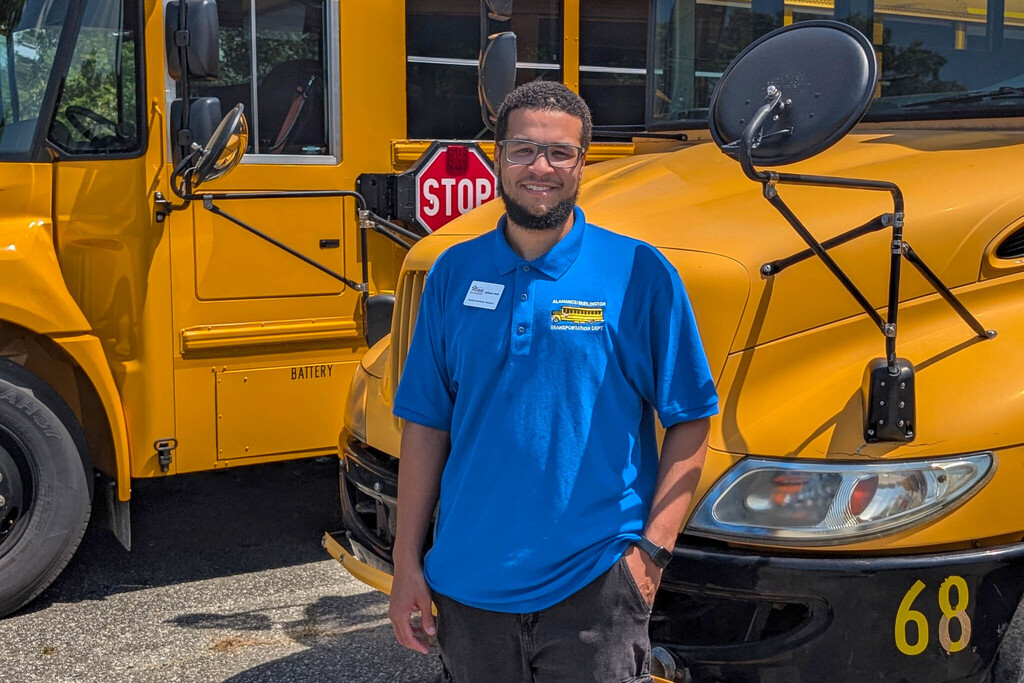 A school bus coordinator in a blue polo smiles while standing in front of two yellow school buses