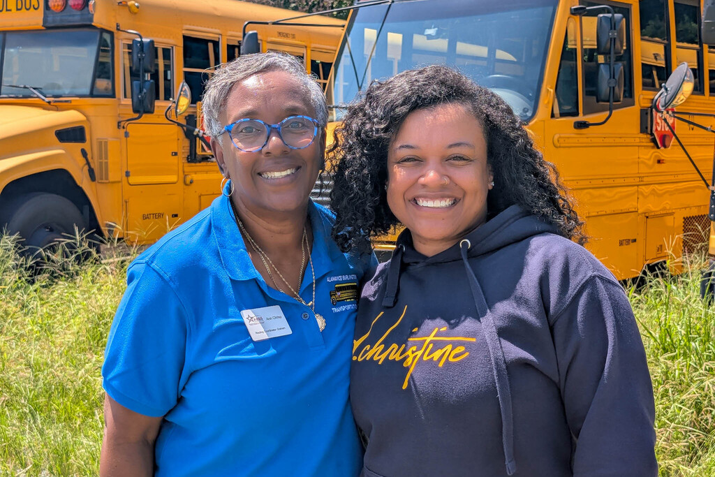 A school bus coordinator in a blue polo stands next to a driver in a blue hoodie in front of yellow school buses and tall grass