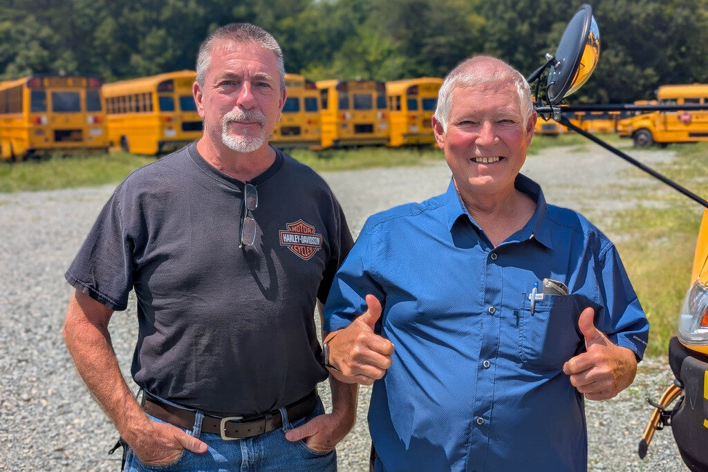 Two bus drivers stand in a gravel road next to many parked school buses.  One is giving a thumbs up.