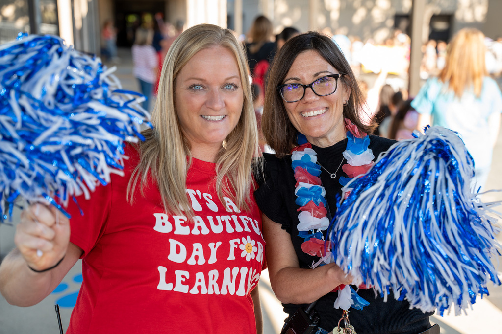 Two school administrators, one wearing black and the other red, wave blue pompoms outside at a first responders celebration event.