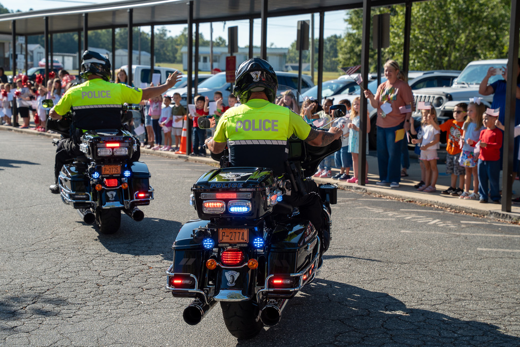 Two police officers on motorcycles drive by and wave at classes of elementary students during a Hero Day Parade event.