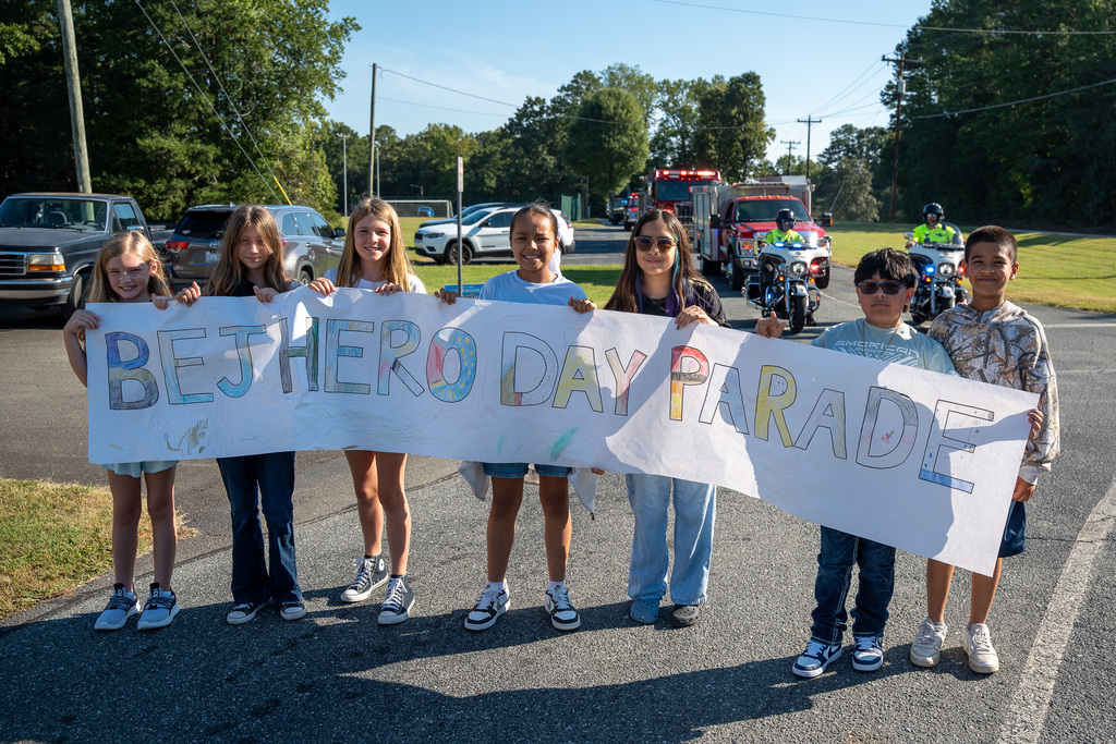 Elementary students stand behind a BEJ Hero Day Parade banner.  Behind them are various first responders in their vehicles, lined up for the parade.