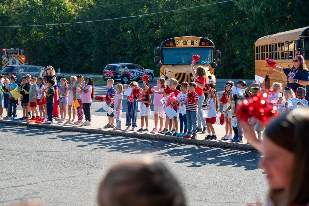 A line of multiple classes of elementary students, most wearing red, white, and blue, and waving pompoms or holding posters, cheer for first responders during a Hero Parade. Behind them are two yellow school buses.