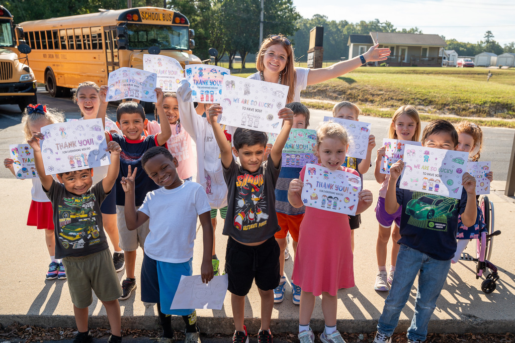 An elementary class hold up thank you signs for first responders with their teacher raising her hand enthusiastically behind them.  In the far background there is a school bus.