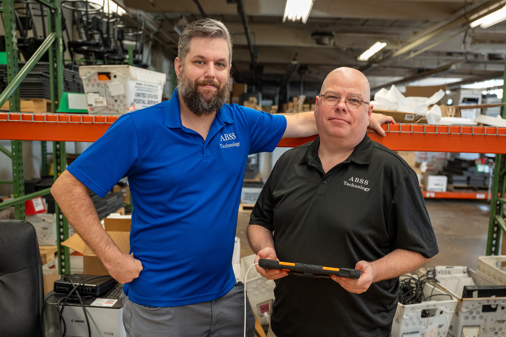 Two technicians, one wearing a blue polo and one wearing a black polo with ABSS Technology embroidery, stand in a warehouse setting, the one in blue propping his hand on a rack behind them