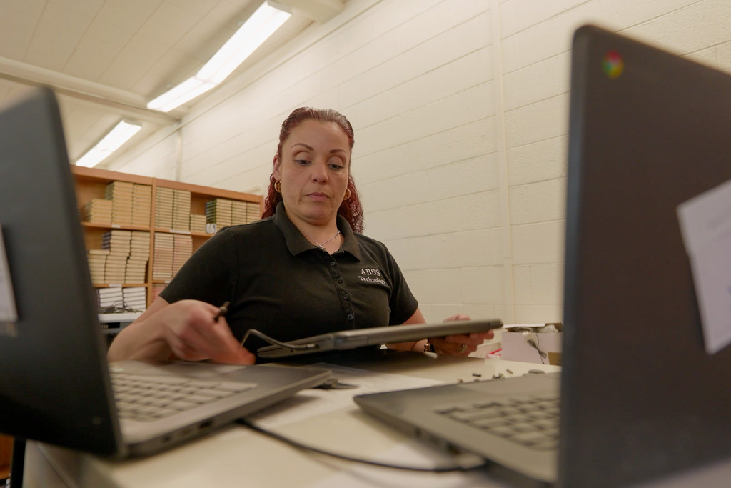A female technician in a black polo pulls a closed Chromebook towards her while addressing a support ticket. Two open Chromebooks are in the foreground.