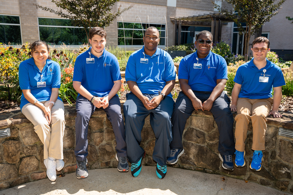 Five high school seniors wearing matching blue Project Search polo shirts are sitting on a rock wall in a hospital courtyard garden