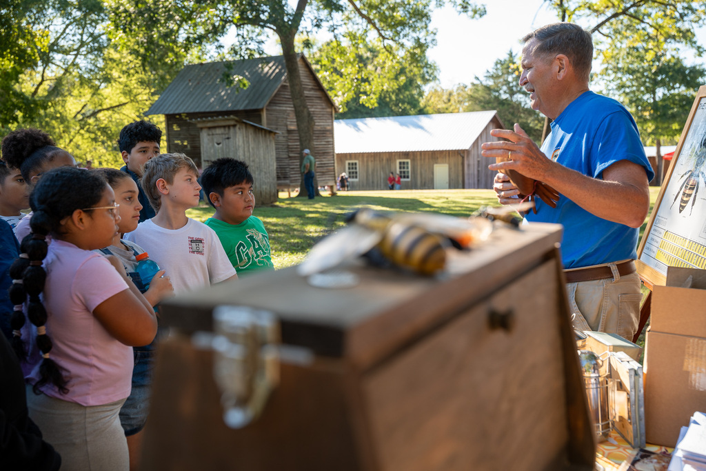 A beekeeper instructs a group of elementary students on bee and beekeeper facts in an outdoor park setting.
