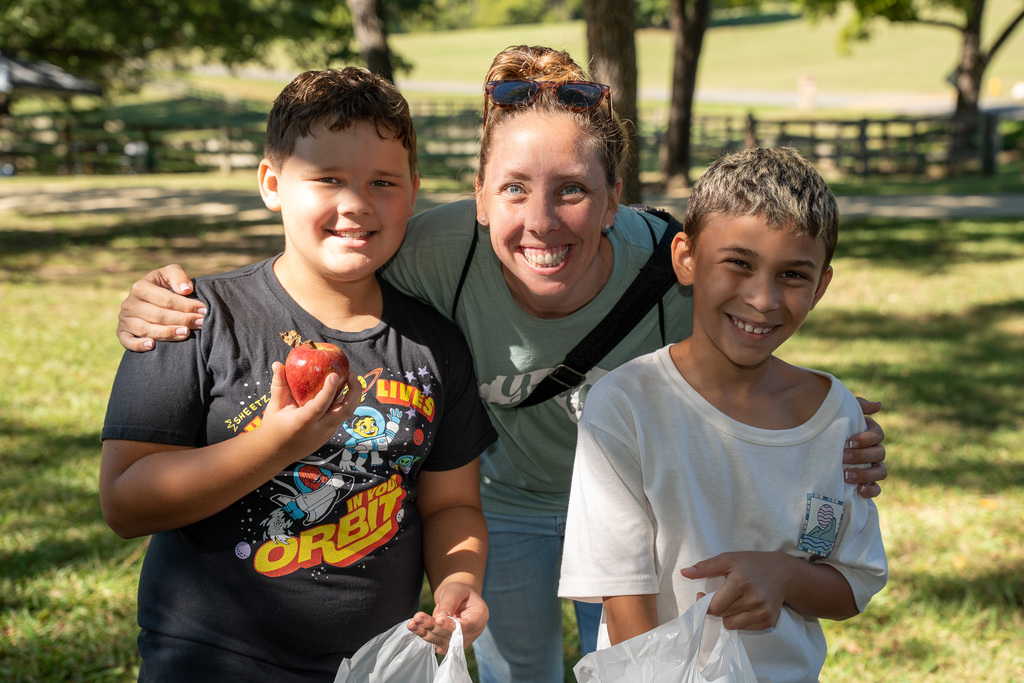 A teacher poses for a photo with two of her students, one is holding an apple, both are holding plastic bags