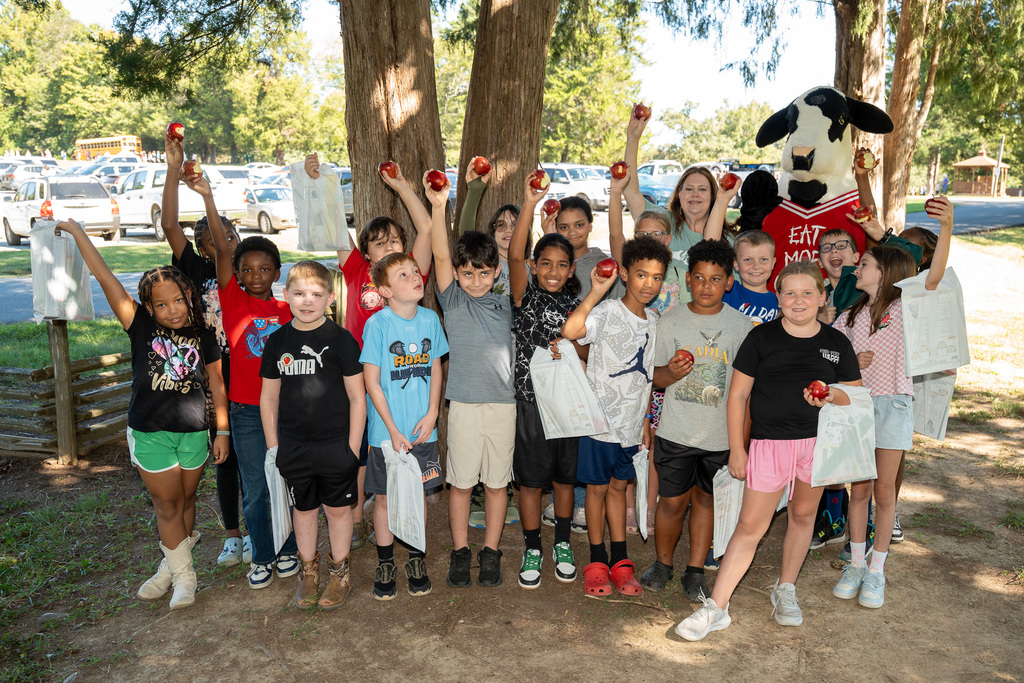 A class of elementary students hold up apples outside in a park next to a Chick-fil-A cow