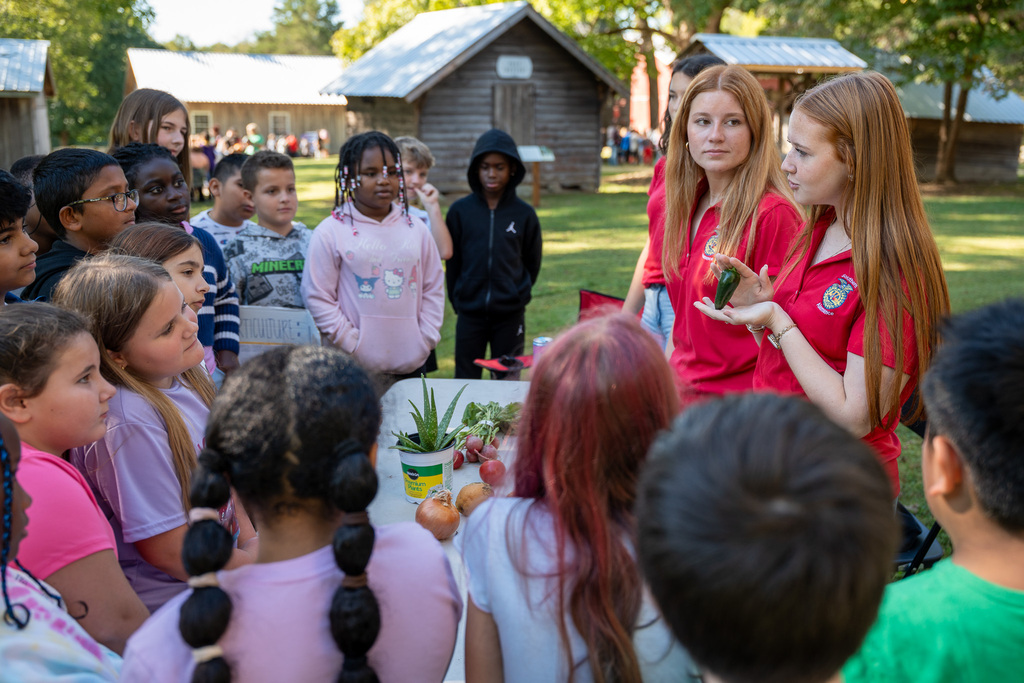 High school FFA students wearing red polo shirts quiz a large group of elementary students on the differences between fruits and vegetables in an outdoor park setting