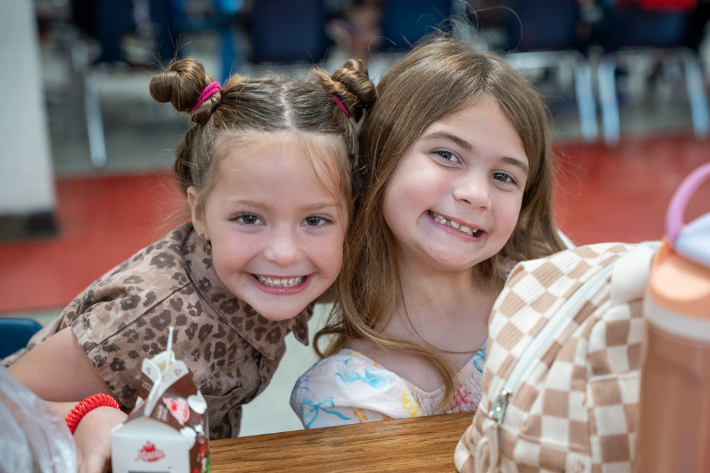 Two young elementary girls smile at a lunch table. A milk carton and lunch bag are in the foreground.