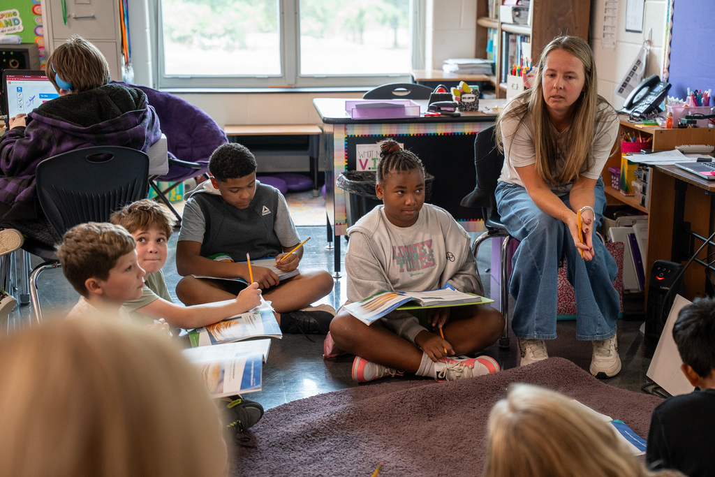 A teacher and elementary students sit around a carpet facing a whiteboard. They are engaged in discussion, with students having workbooks in their laps.