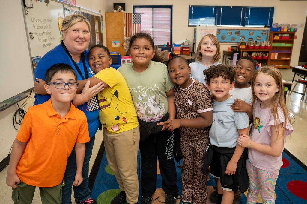 A teacher stands beside eight of her students, with one of them giving a side hug.