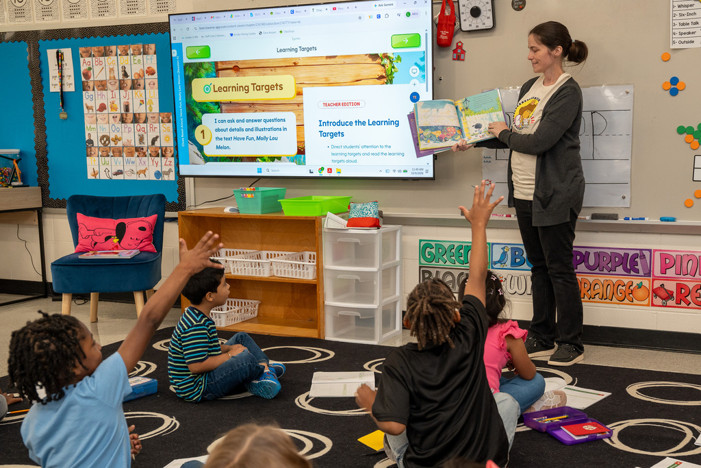 A teacher stands at the front of the room reading a book with an attentive group of young elementary students on the carpet. Two of them have their hands held high.
