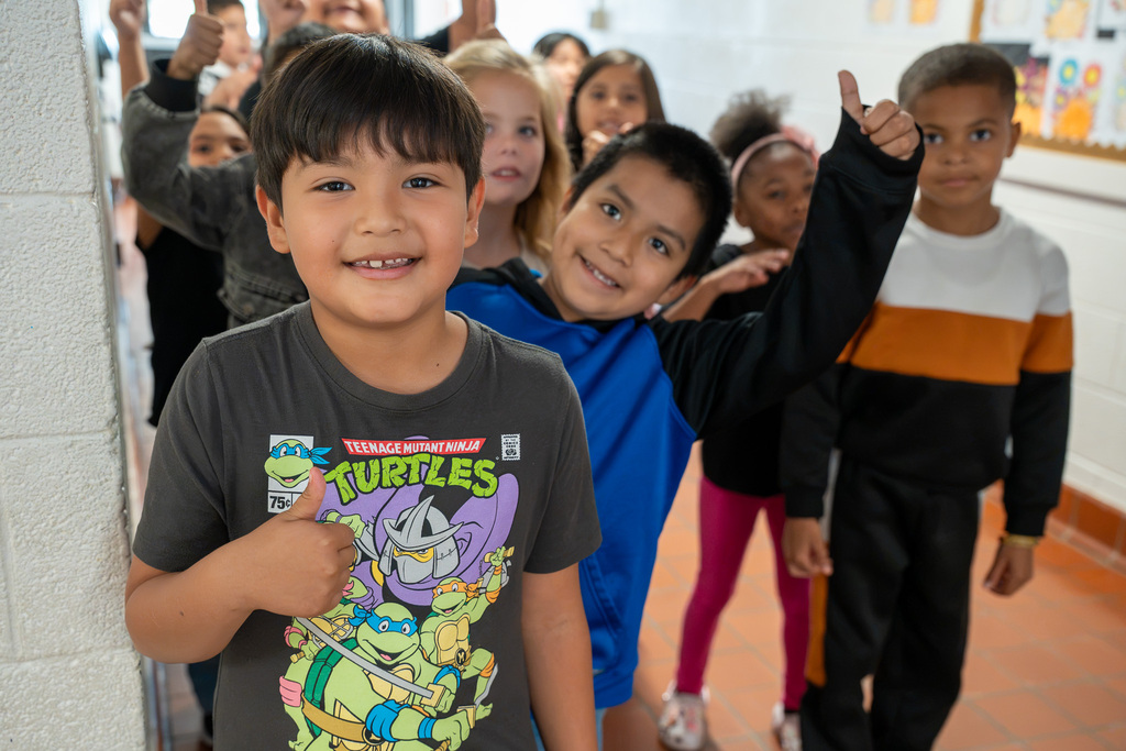 A class of elementary students in the hallway looks towards the camera, with a couple of them smiling and giving a thumbs up.