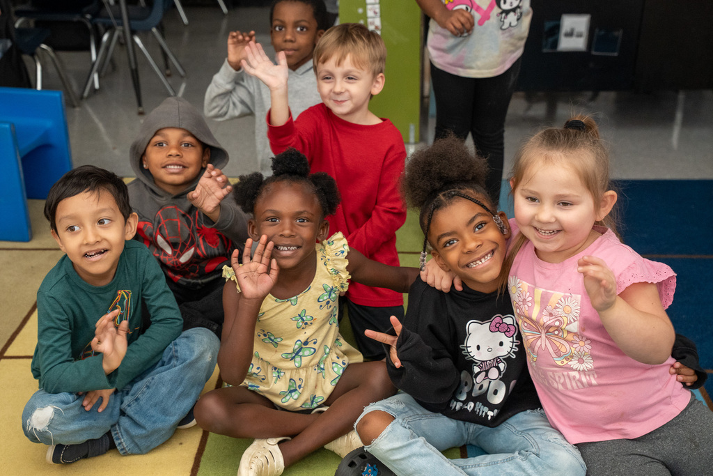 A diverse class of young elementary students waves and smiles as they are seated on a classroom carpet
