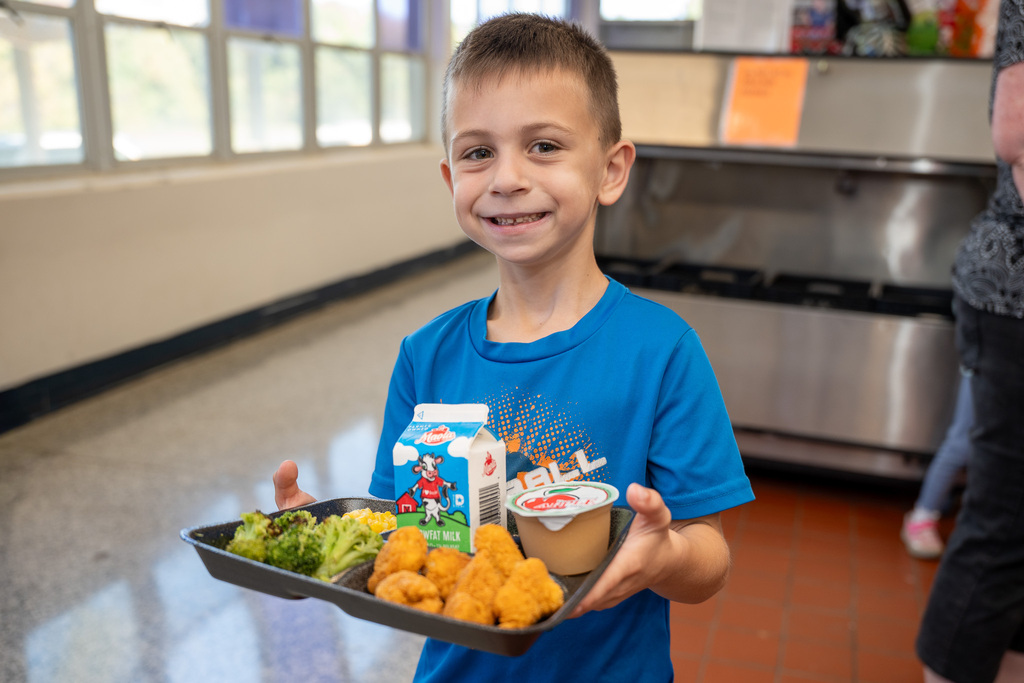 A young boy wearing a blue t-shirt holds a lunch tray with milk, chicken bites, broccoli, and apple sauce.