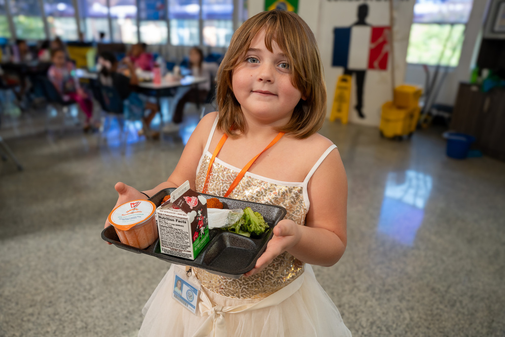 A young girl holds a lunch tray with broccoli, chicken bites, a fruit cup, and chocolate milk