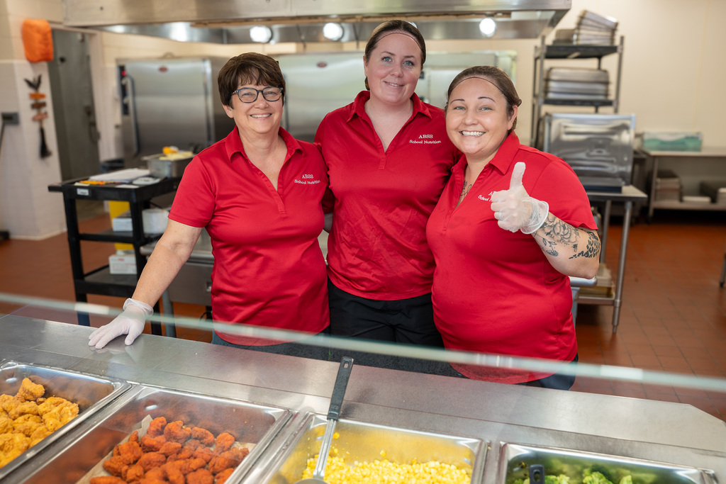 School nutrition staff wearing red shirts pose for a photo behind the serving line.  Various school foods are seen in the foreground, including chicken bites, corn, and broccoli.