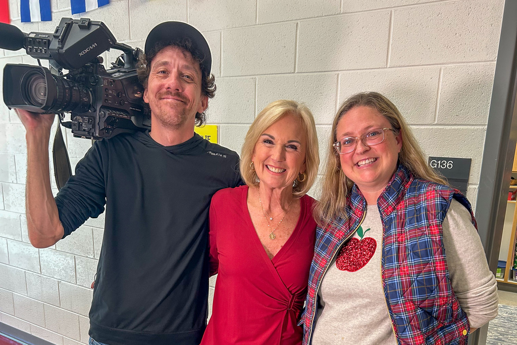 A cameraman with a shoulder-mounted TV camera, a news reporter in a red top, and teacher Brandy Brehm smile together in a school hallway.
