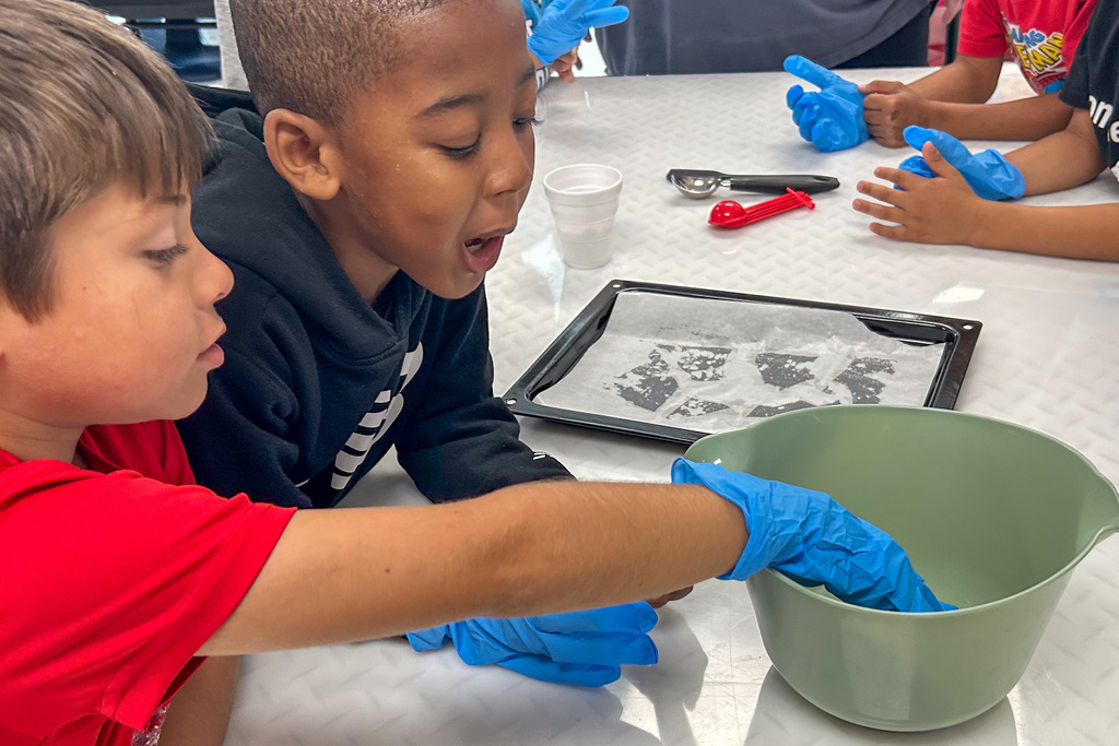 Two excited students wearing blue gloves reach into a green mixing bowl beside a baking tray at a classroom table, while other students look on.
