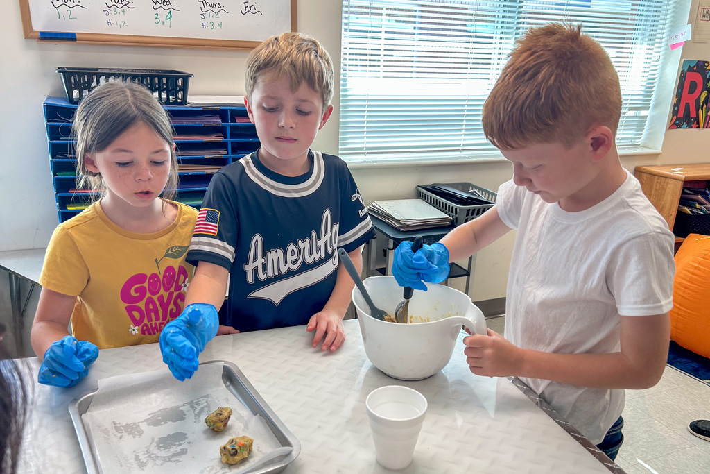 Three elementary students wearing blue gloves mix cookie dough in a white bowl and place dough on a baking tray at a classroom table.