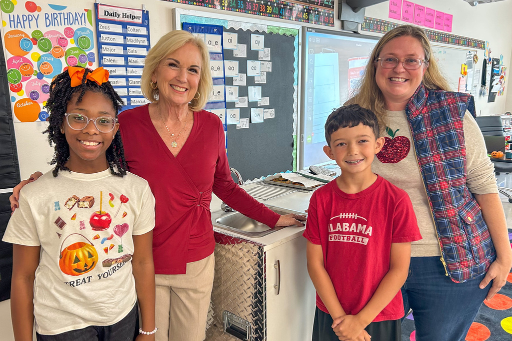 A reporter, teacher Brandy Brehm, and two smiling students stand beside the new mobile kitchen, which has a sink and a diamond-plate metal side.