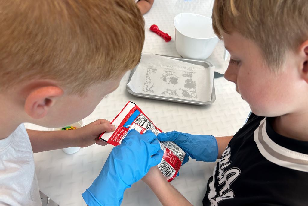 Two students in blue gloves read the cookie dough package directions together, with a baking tray and mixing bowl on the table behind them.