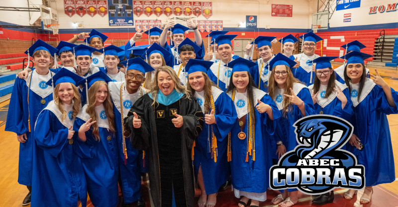 Group photograph of high school graduates and principal of Early College at ACC wearing blue robes, giving thumbs up, in a school gynasium