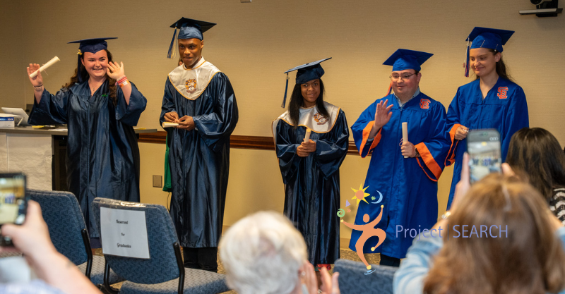 Five high school graduates wearing blue caps and gowns from different schools celebrating in front of a crowd
