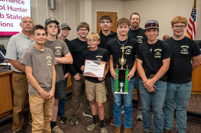 The Southern Middle Hunter Safety team wearing mostly matching black or gray shirts, some have hats, the student in the front is holding a trophy and the student beside him is holding a certificate
