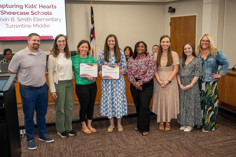 A mix of principals and educators, two holding certificates of recognition, at the board of education meeting during recognitions