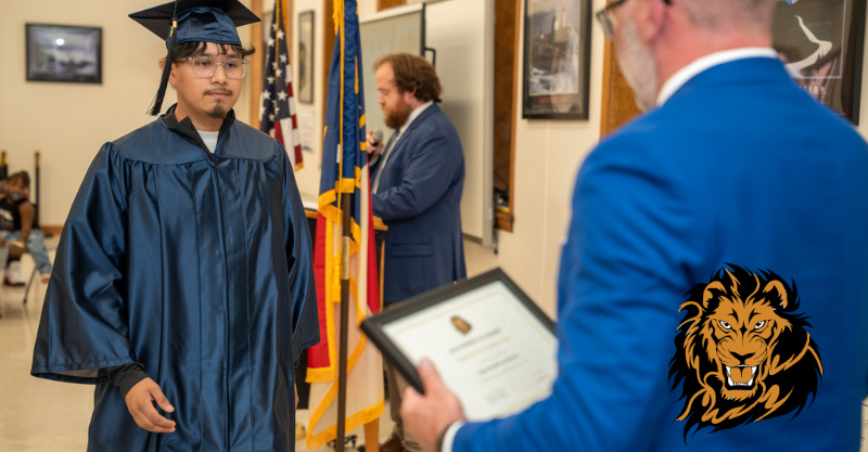 A high school graduate in blue robes walks up to accept a certificate at a ceremony from the principal in a blue suit