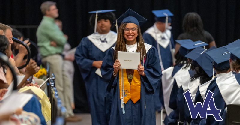 A high school graduate wearing blue robes with a yellow sash holds up a white certificate holder as she proceeds down a center aisle
