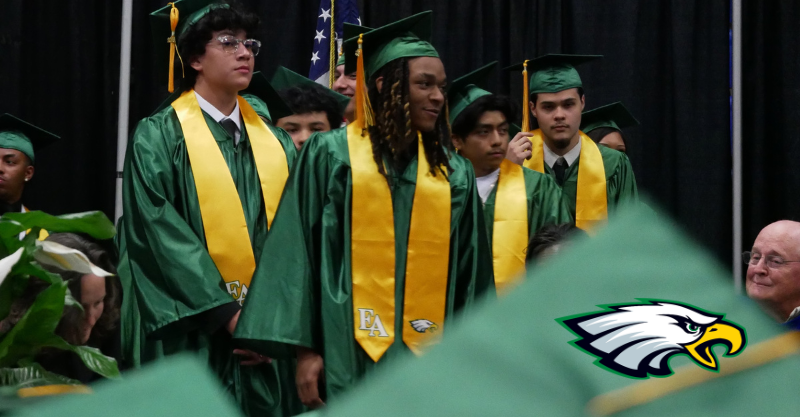 High school seniors wearing green and yellow format graduation attire standing on stage ready to walk across to receive their diploma